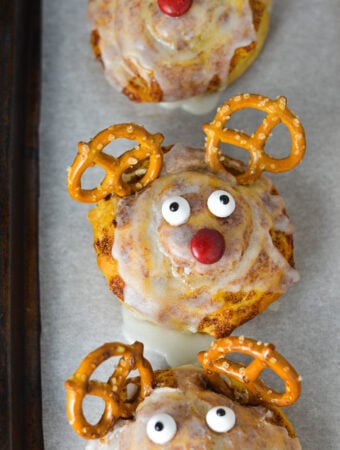 A parchment paper lined cookie sheet with Easy Reindeer Cinnamon Rolls on top: premade cinnamon rolls made to look like Rudolph the red nosed reindeer.