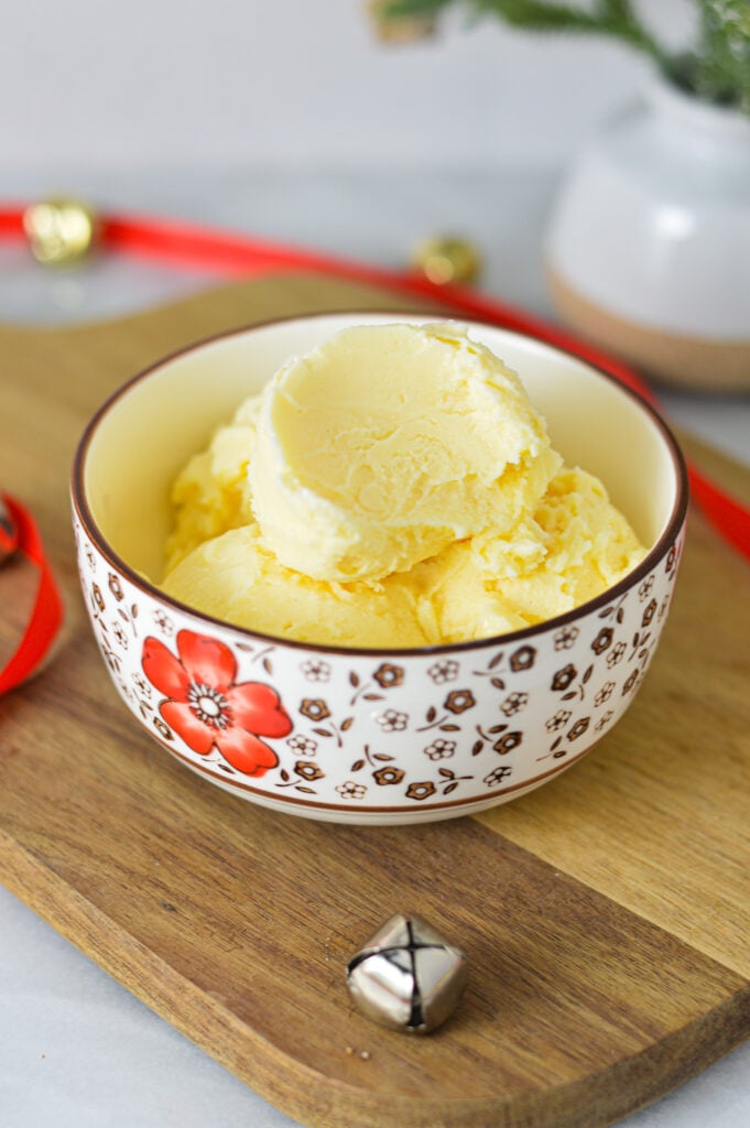 A small bowl filled with Ninja Creami Eggnog Ice Cream, with red ribbon and gold bells in the background giving it a holiday season feel.
