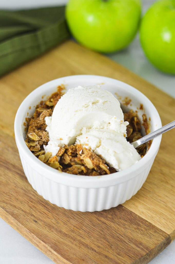 A small spoon in an individual apple crisp, with the vanilla ice cream melting slightly in the crisp topping.
