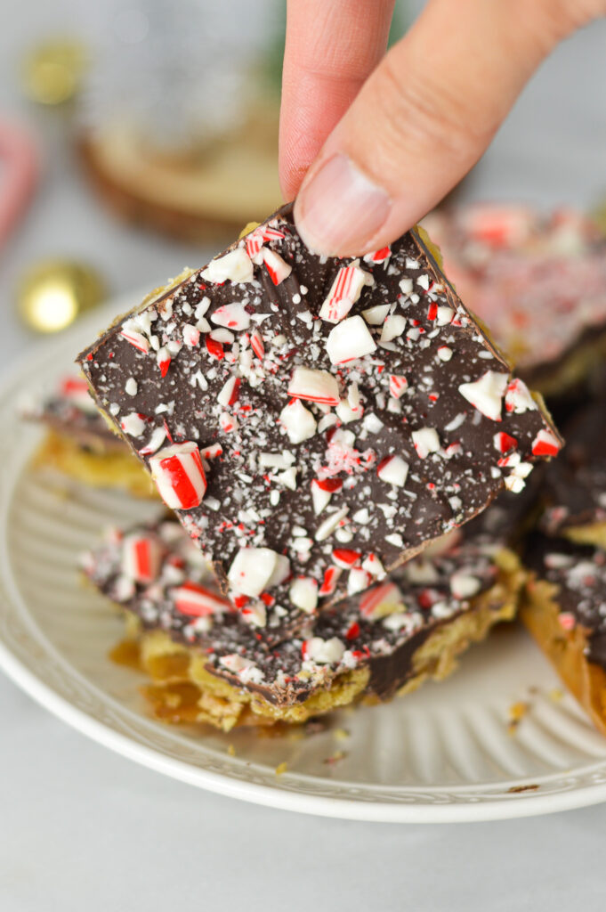 A hand holding up a piece of Candy Cane Saltine Toffee, with gold bells and whole candy canes in the background, giving it a Christmas feel.