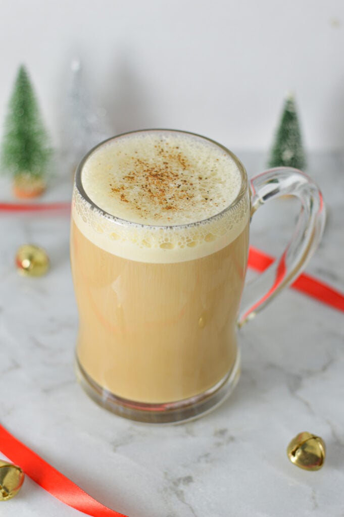 Eggnog Latte in a glass mug, with some red ribbon and gold bells in the background giving it a Christmas feel.