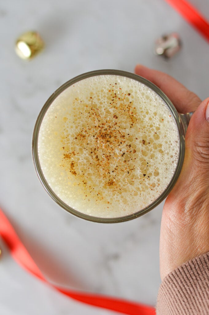 A hand warming up with a mug of Eggnog Latte, with some Christmas bells and ribbon on the granite surface.