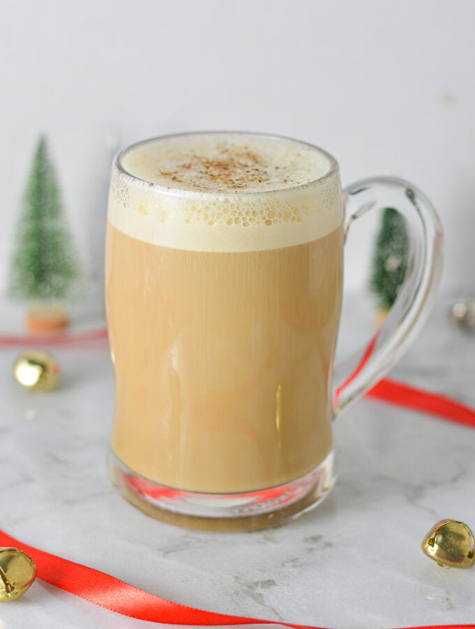 A clear mug full of Eggnog Latte, with the frothy milk topped with ground nutmeg.