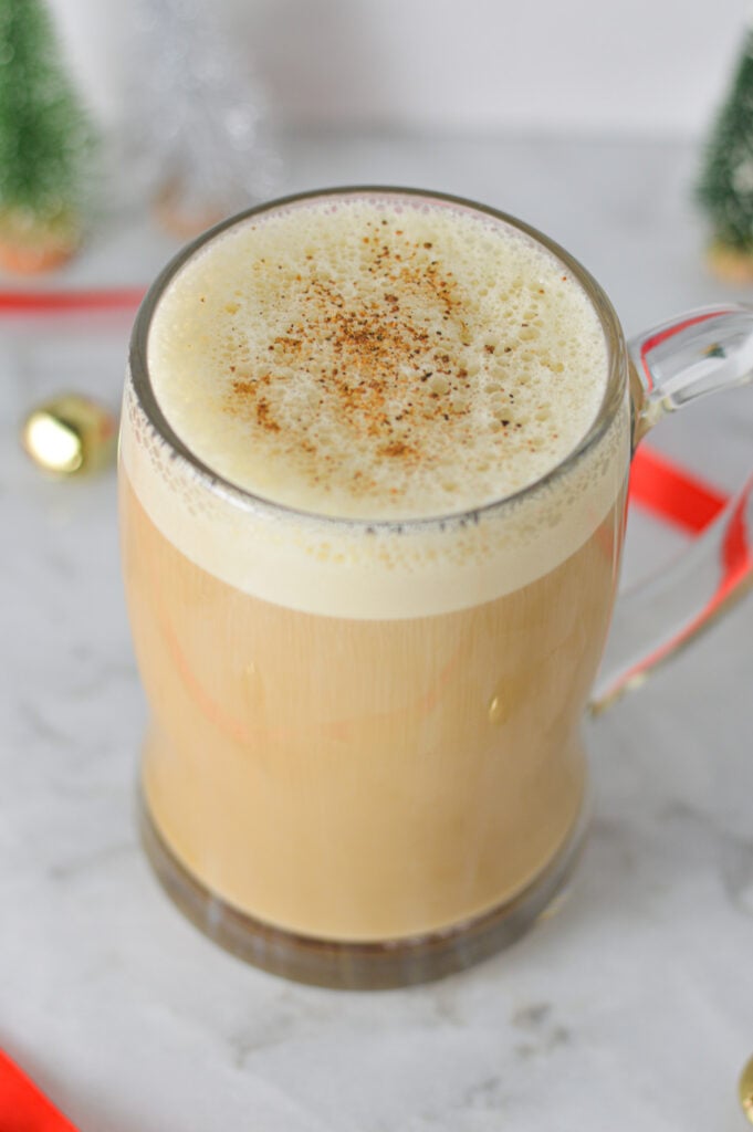 A large mug full of Eggnog Latte, with some golden bells and wrapping ribbon in the background giving it a holiday season vibe.