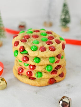 A stack of Mini M&M Christmas Cookies with red ribbon and silver and gold bells in the background, giving the scene a real holiday season feeling.