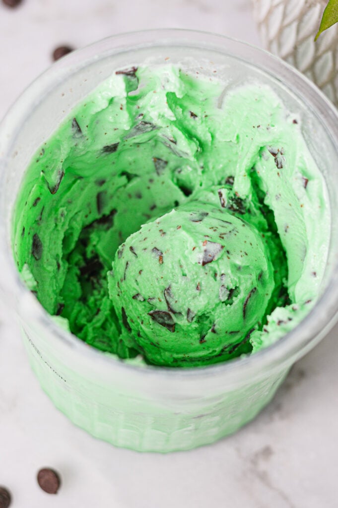A Ninja Creami pint container filled with vibrant green Ninja Creami Mint Chocolate Chunk Ice Cream, with a scoop showing the chocolate filled creamy ice cream.