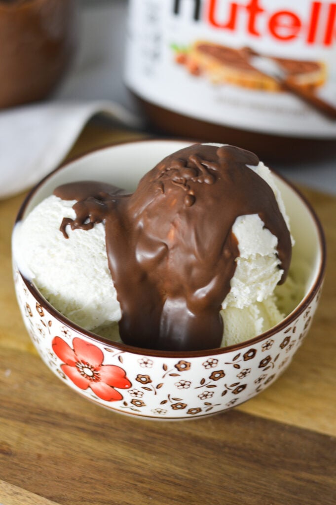 A bowl of vanilla ice cream topped with Nutella Magic Shell, with a jar of Nutella in the background.