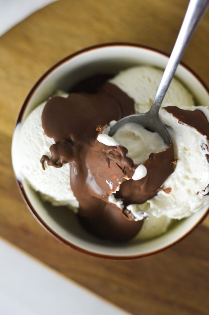 A bowl full of vanilla bean ice cream, topped with Nutella Magic Shell. A spoon is taking a scoop of ice cream, cracking the hard chocolate shell.