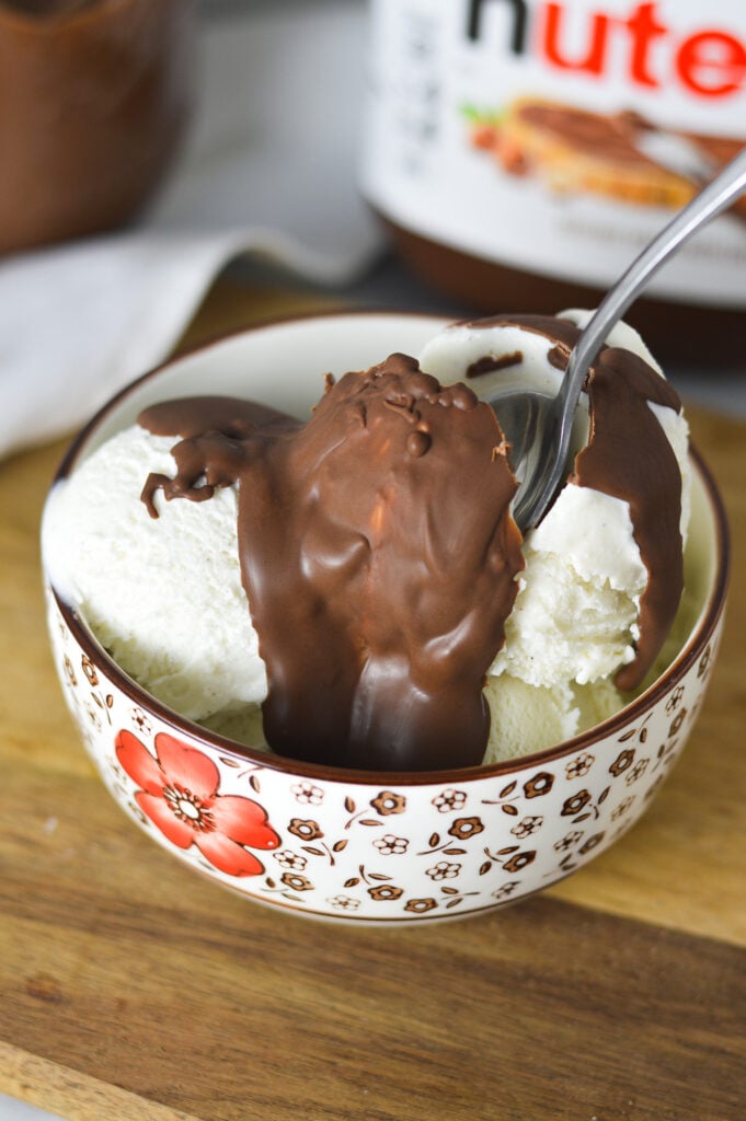 A small spoon inserted into a small bowl full of ice cream topped with Nutella Magic Shell. The spoon is cracking the hard shell of chocolate.