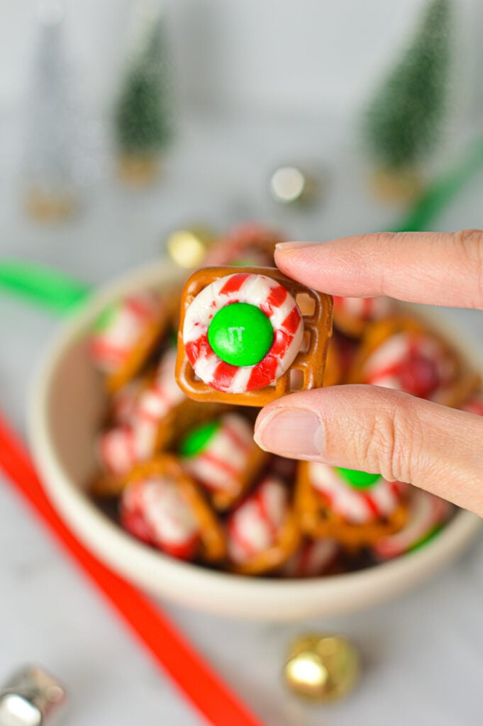 A hand holding a Peppermint Kiss Pretzel, with a bowl full of the festive treats in the background.