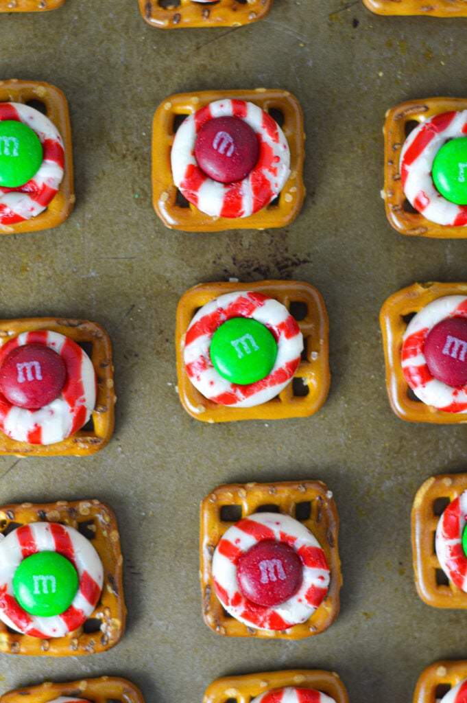 Some homemade Peppermint Kiss Pretzels on a cookie sheet, showing the candy cane kisses, square pretzels and holiday M&Ms clearly.