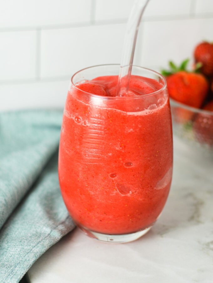 A glass of Virgin Frozen Strawberry Daiquiri on a granite surface, with fresh strawberries in the background giving the drink a refreshingly fresh feel.