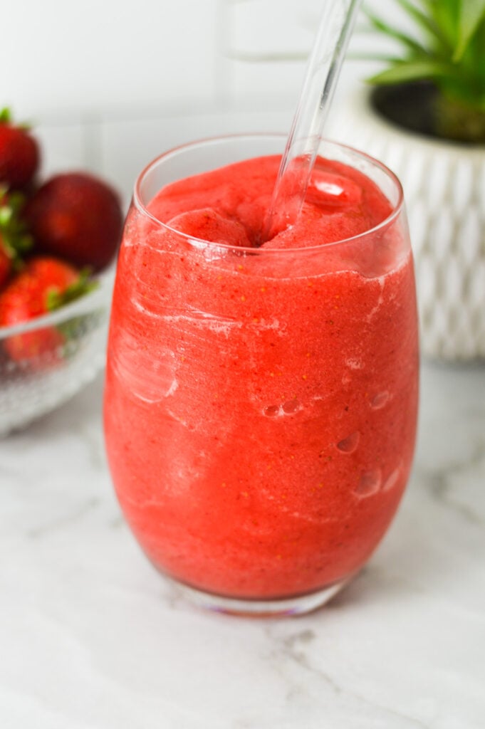 A glass of Virgin Frozen Strawberry Daiquiri with a small bowl full of bright red strawberries, and a straw sticking out of the virgin drink.