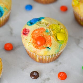 Freshly baked M&M Muffins on a granite surface, with some chocolate M&Ms surrounding the soft muffins.