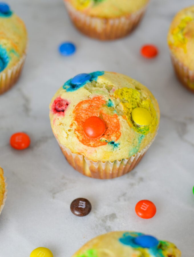 Freshly baked M&M Muffins on a granite surface, with some chocolate M&Ms surrounding the soft muffins.