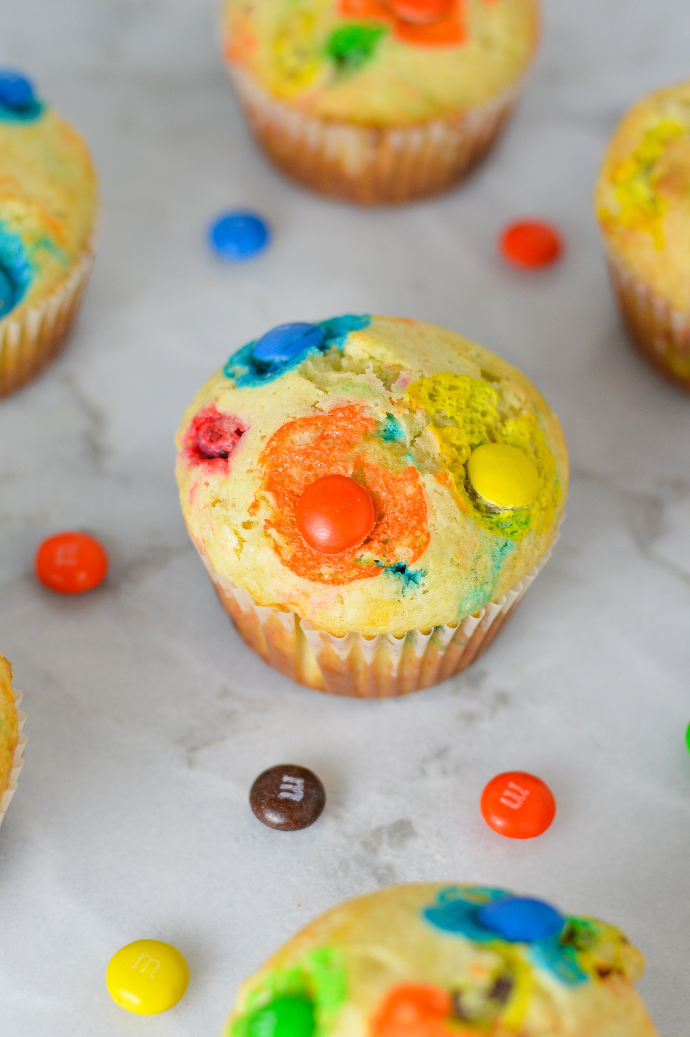 Freshly baked M&M Muffins on a granite surface, with some chocolate M&Ms surrounding the soft muffins.