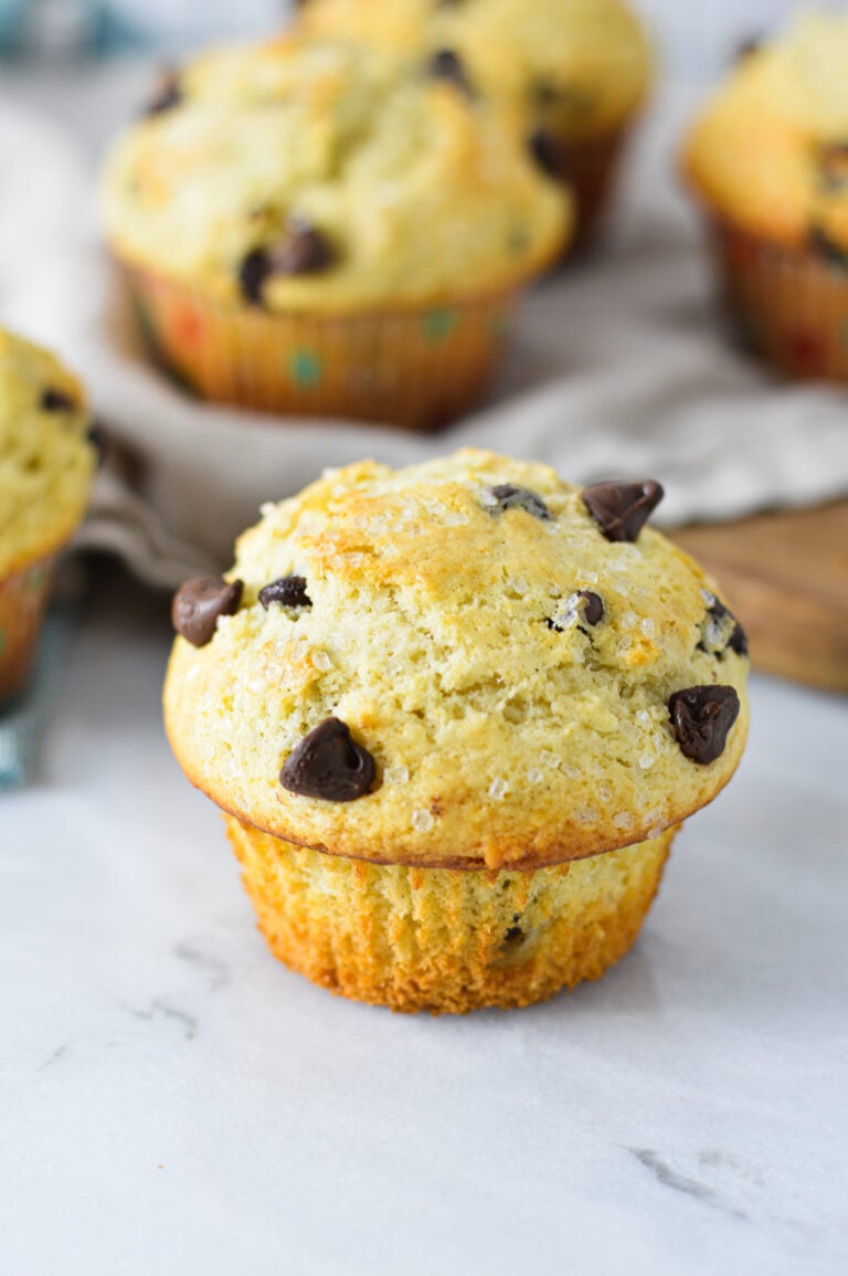 Some fluffy Chocolate Chip Muffins on a granite surface, with the paper liners taken off of the front muffin.