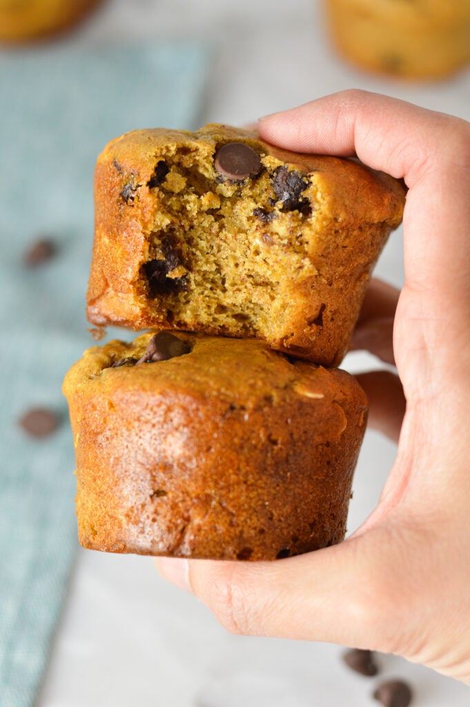 A hand holding a stack of Healthy Peanut Butter Chocolate Chip Oat Muffins, with a bite taken out of the top muffin showing the fluffy inside.