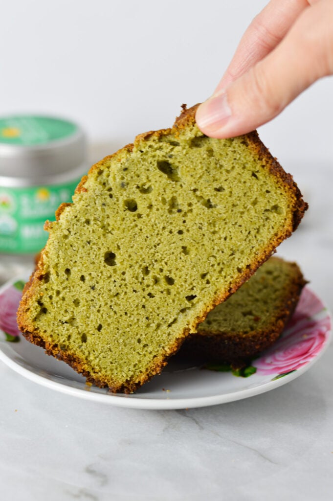 A hand taking a slice of Matcha Pound Cake off of a small plate, with a jar of matcha powder in the background.