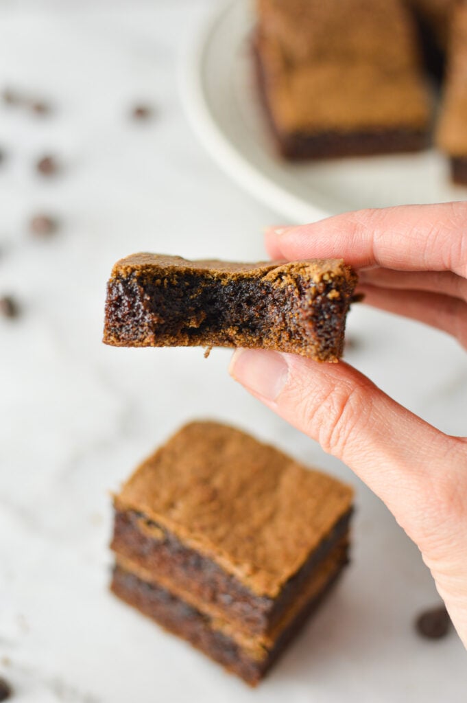 A hand holding a Mayonnaise Brownie with a bite taken out, showing the fudgy texture of the inside of the brownie.