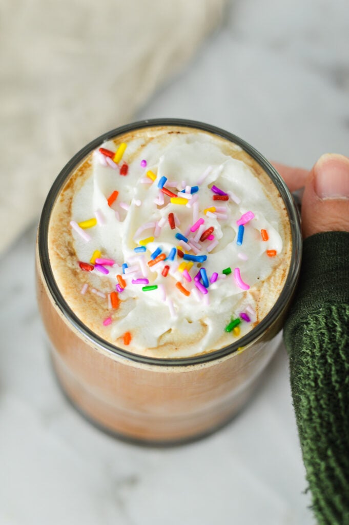A hand holding a coffee mug full of homemade Cake Batter Mocha, topped with whipped topping and colorful sprinkles.