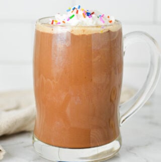 A tall glass mug full of rich Cake Batter Mocha, topped with homemade whipped cream and multicolored sprinkles.