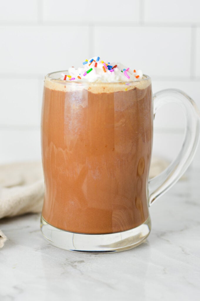 A tall glass mug full of rich Cake Batter Mocha, topped with homemade whipped cream and multicolored sprinkles.