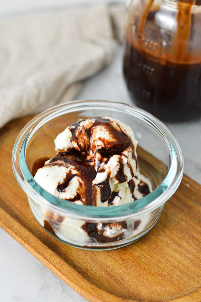 A small bowl filled with vanilla ice cream topped with Homemade Chocolate Sauce, with a jar of the homemade sauce in the background.