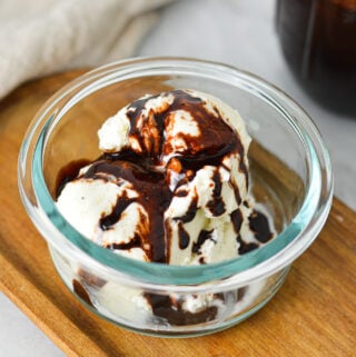 A small glass bowl filled with scoops of ice cream topped with Homemade Chocolate Sauce, with a glass jar of the quick chocolate sauce in the background.