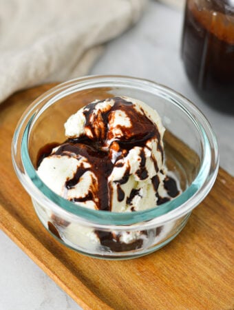 A small glass bowl filled with scoops of ice cream topped with Homemade Chocolate Sauce, with a glass jar of the quick chocolate sauce in the background.