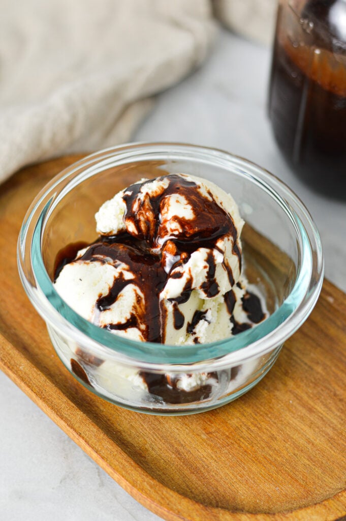 A small glass bowl filled with scoops of ice cream topped with Homemade Chocolate Sauce, with a glass jar of the quick chocolate sauce in the background.