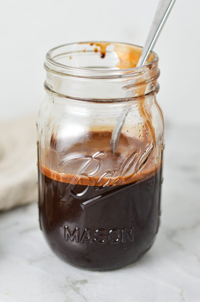 A medium mason jar filled with Homemade Chocolate Sauce, with a spoon in the sticky sauce showing the pourable texture.