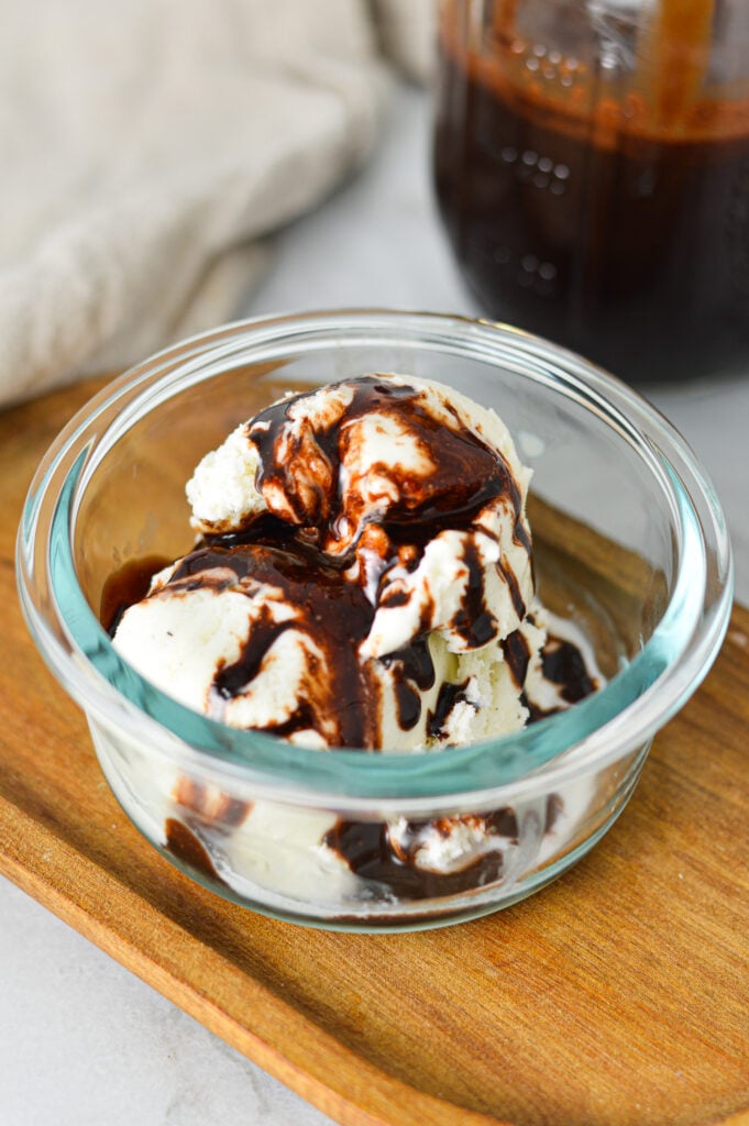Scoops of vanilla bean ice cream in a glass bowl with some chocolate drizzle on top from the Homemade Chocolate Sauce in the mason jar in the background.