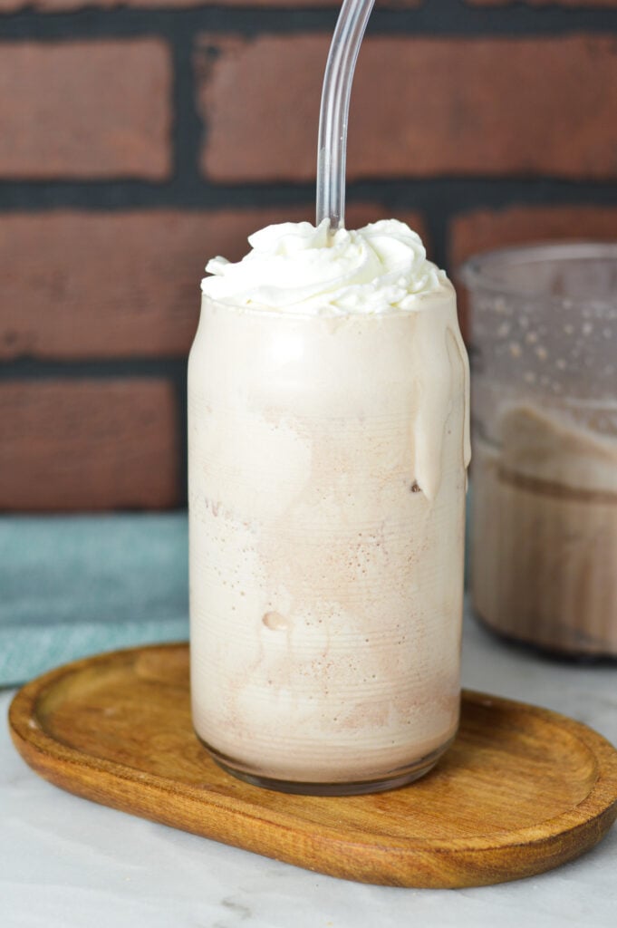 A large glass filled with Ninja Creami Frozen Hot Chocolate, with a straw sticking out of the dessert drink and a Ninja Creami container filled with leftover drink in the background.