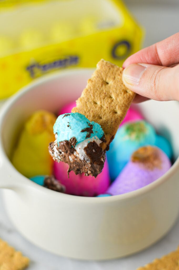 A piece of graham cracker cookie taking a scoop of Peeps S'mores Dip from a small oven safe container.