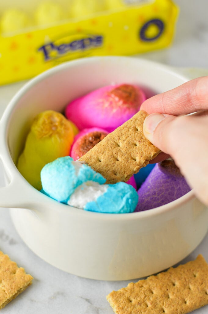 A graham cracker cookie taking a scoop of Peeps S'mores Dip from a small container, with the melty Easter marshmallows breaking up on top of the melted chocolate.