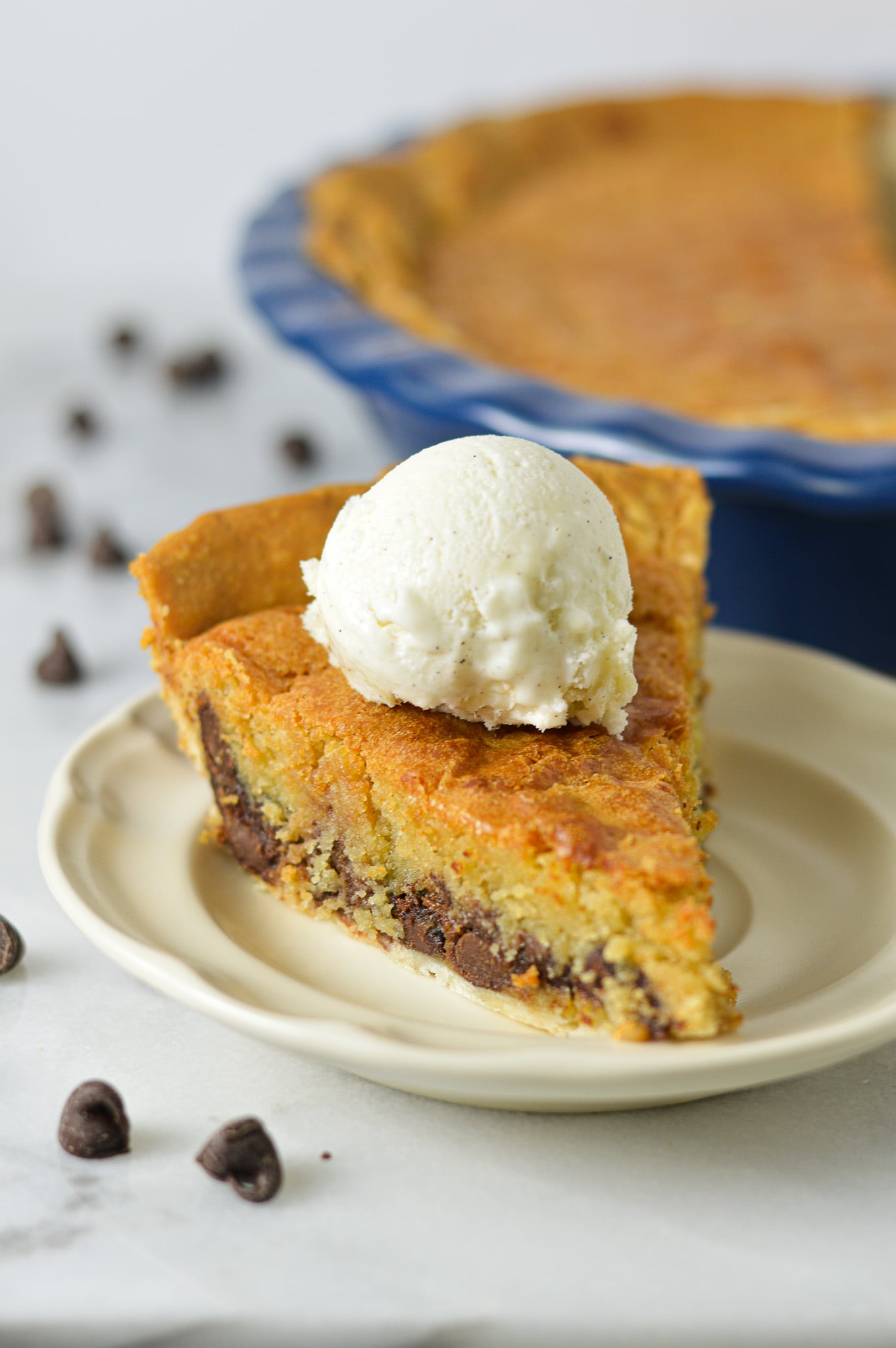 A slice of Chocolate Chip Cookie Pie on a small plate, with the whole pie in its ceramic pie pan in the background.