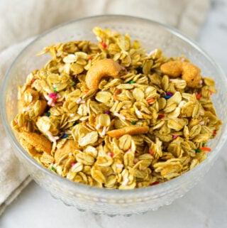 A glass bowl full of freshly baked Funfetti Granola, with almond pieces and rainbow sprinkles among the granola clusters.