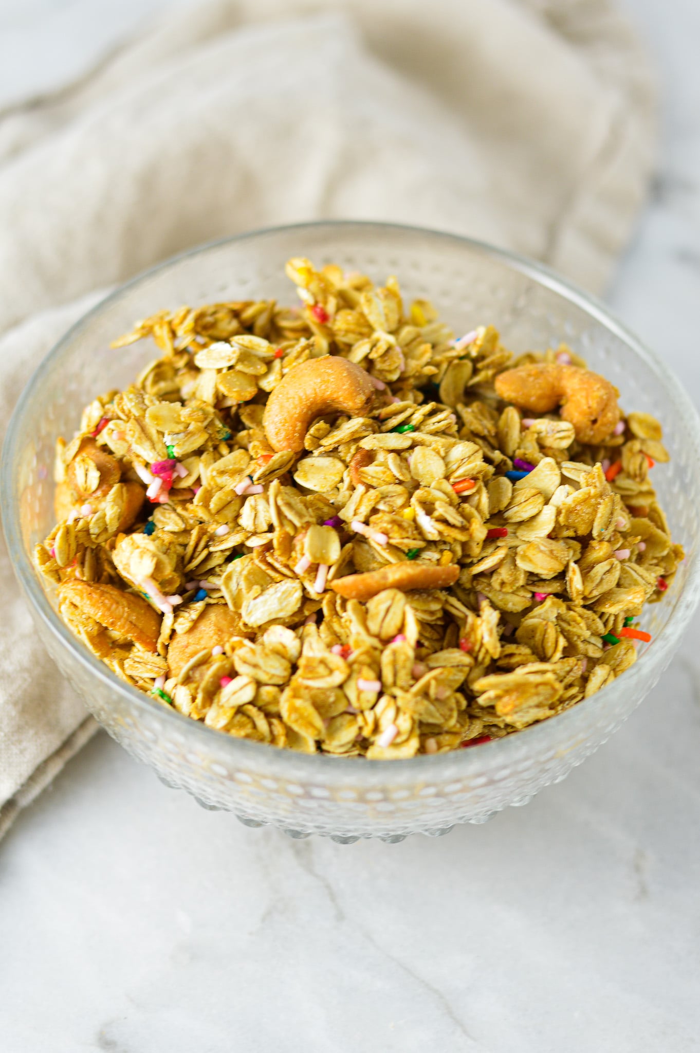 A glass bowl full of freshly baked Funfetti Granola, with almond pieces and rainbow sprinkles among the granola clusters.
