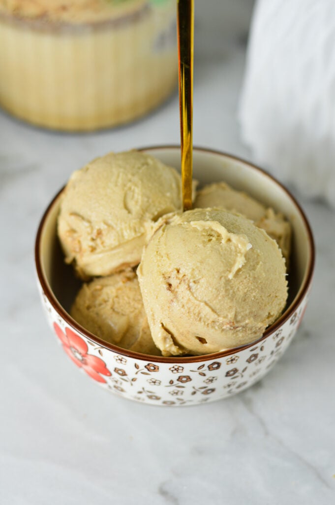 A small bowl filled with homemade Ninja Creami Cinnamon Protein Ice Cream, with a Ninja Creami pint in the background full of leftover ice cream.