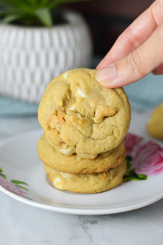 A small plate with a stack of White Chocolate Cashew Cookies. A hand is taking the first homemade cookie, showing the golden brown cookie.