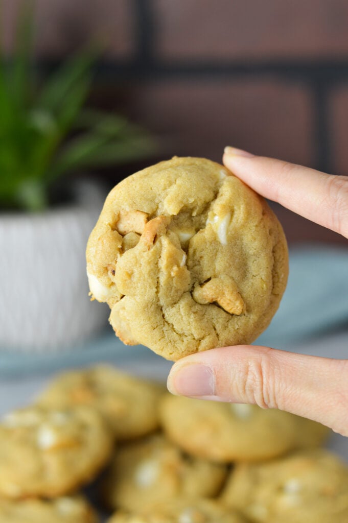 A hand holding up a White Chocolate Cashew Cookie, witht a plate full of the chewy cookies in the background.