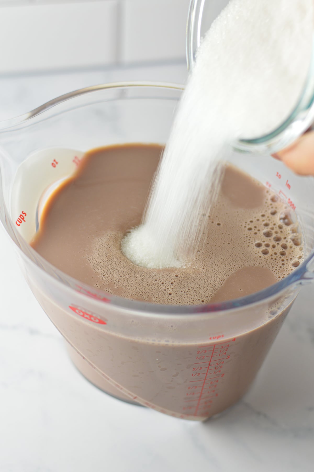 White sugar being poured into a large glass measuring cup full of chocolate milk.