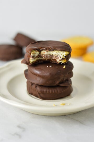 Stack of three chocolate-covered Nutella Ritz cracker sandwiches on a white plate, with a bite taken out of the top cracker showing the Nutella filling.