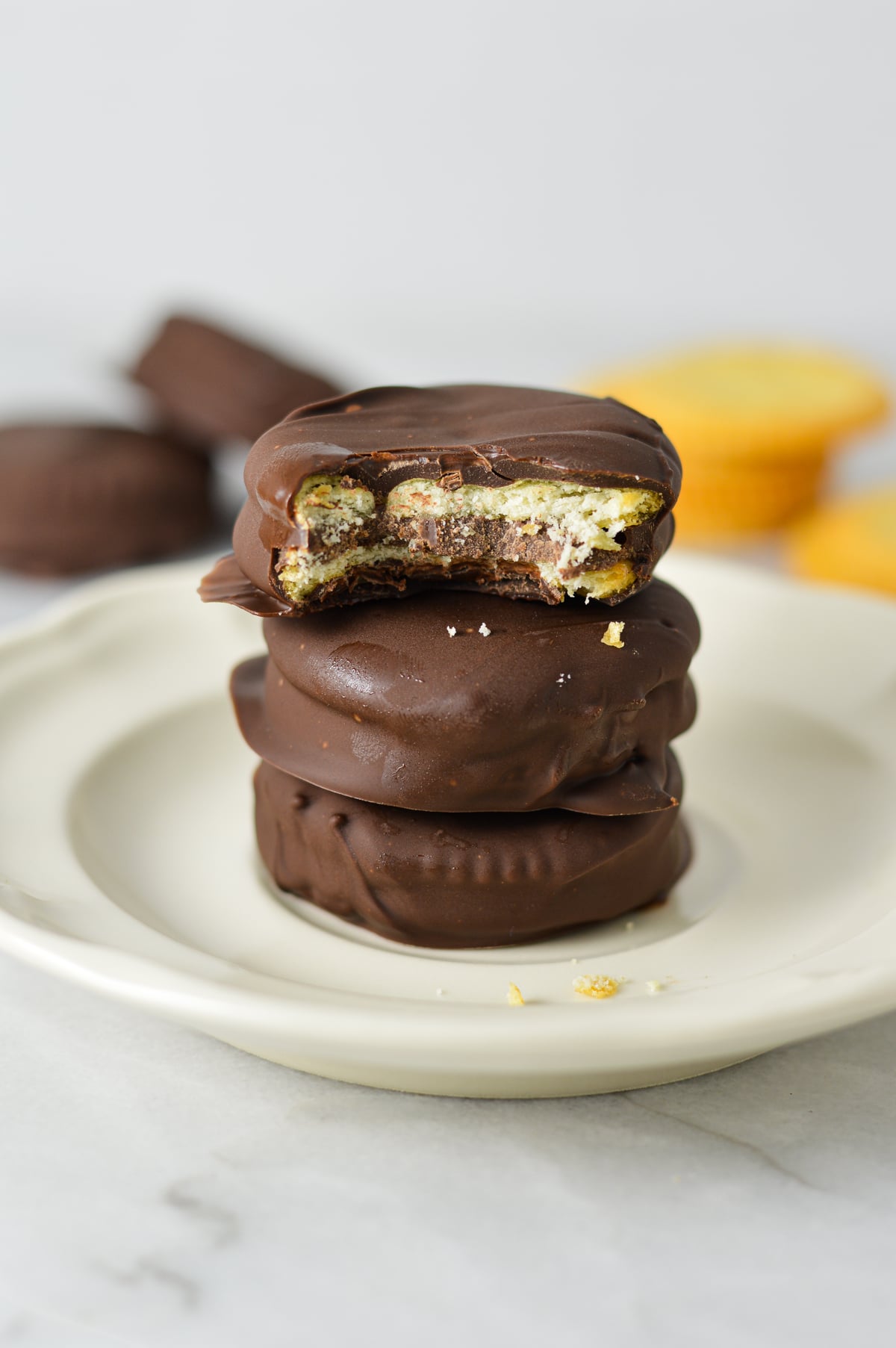 Stack of three chocolate-covered Nutella Ritz cracker sandwiches on a white plate, with a bite taken out of the top cracker showing the Nutella filling.
