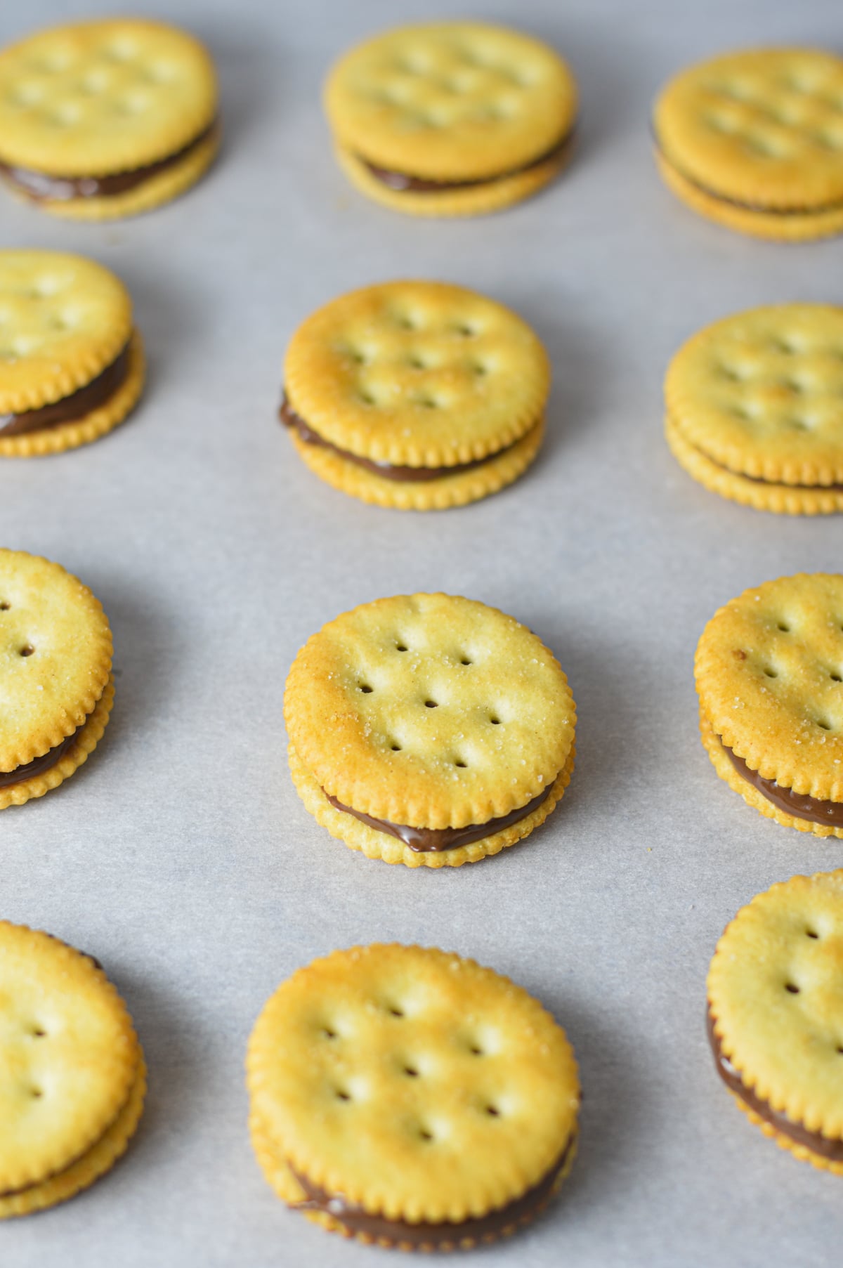 Ritz cracker sandwiches spread with Nutella arranged in rows on a parchment paper-lined baking sheet.
