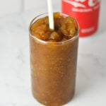 A tall glass full of Ninja Slushi Rum and Coke, with a plastic straw in the slushie drink and a can of Coke in the background.