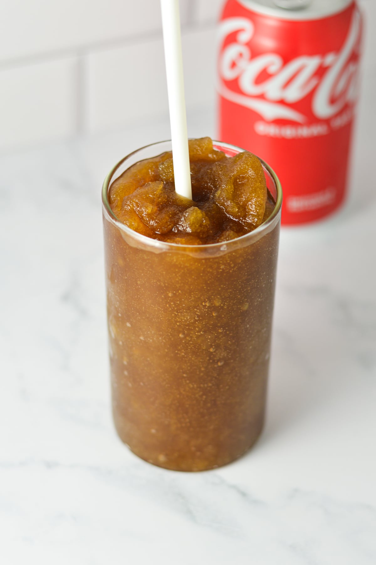 A tall glass full of Ninja Slushi Rum and Coke, with a plastic straw in the slushie drink and a can of Coke in the background.