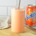 A tall glass filled with a slushy orange creamsicle drink, next to a can of Sunkist Orange soda on a kitchen counter.