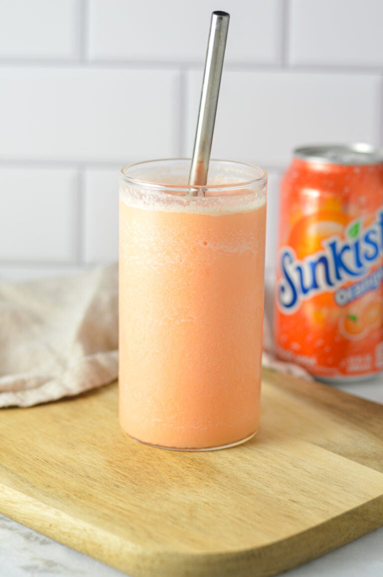 A tall glass filled with a slushy orange creamsicle drink, next to a can of Sunkist Orange soda on a kitchen counter.